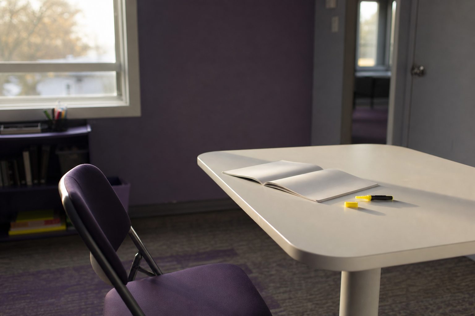An empty chair pulled back from a small white tutoring table with an open notebook and uncapped yellow highlighter