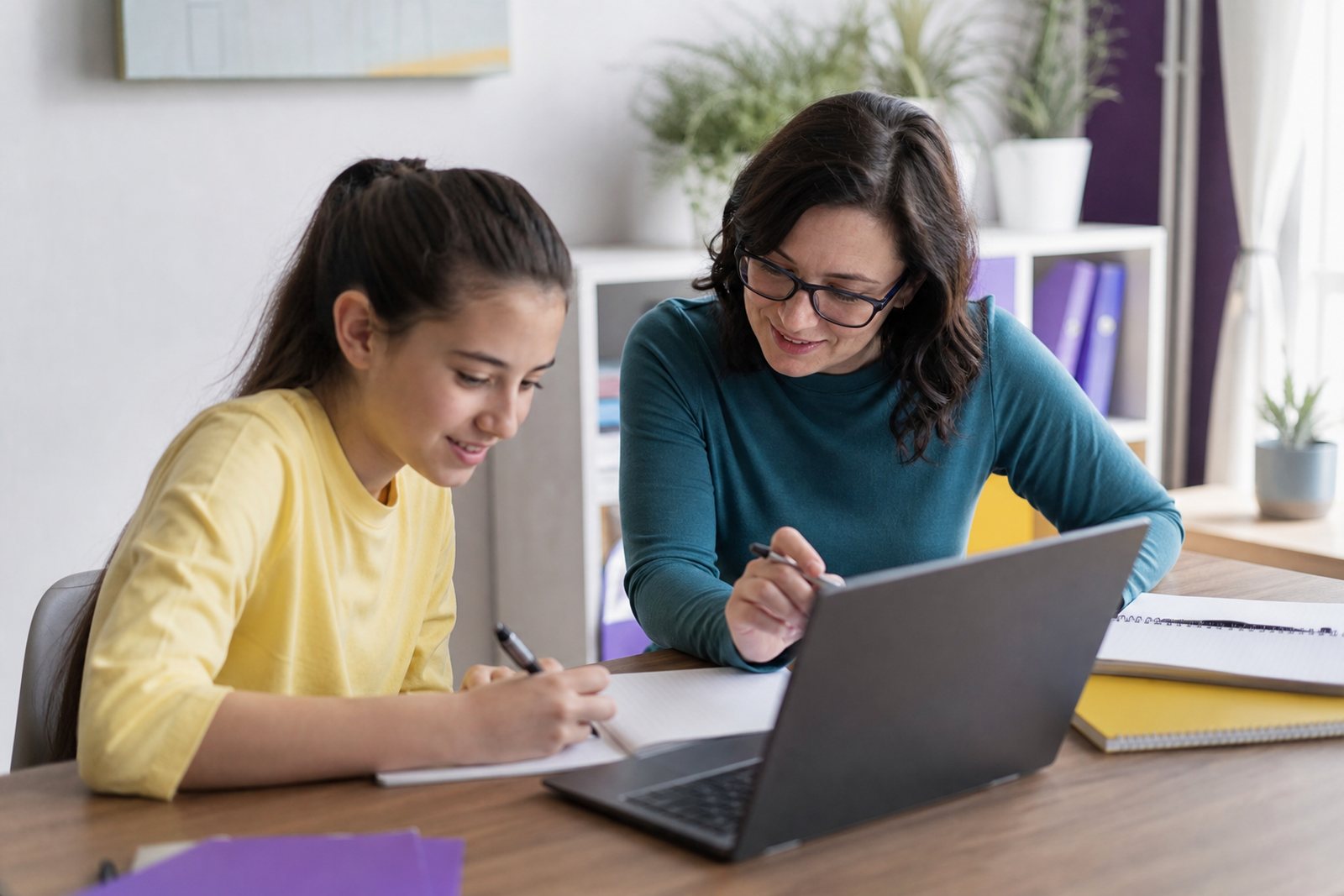 A tutor sitting beside a middle school student at a bright study table covered with notebooks and a laptop